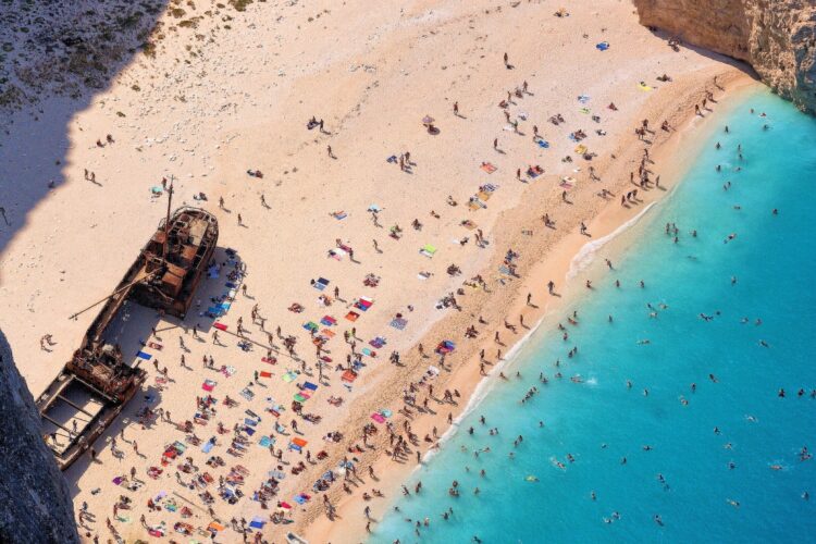 ludzie widziani z lotu ptaka na plaży oblanej turkusową wodą z wrakiem statku u wybrzeża