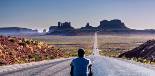 chłopak siedzi na środku drogi patrząc na monument valley w usa