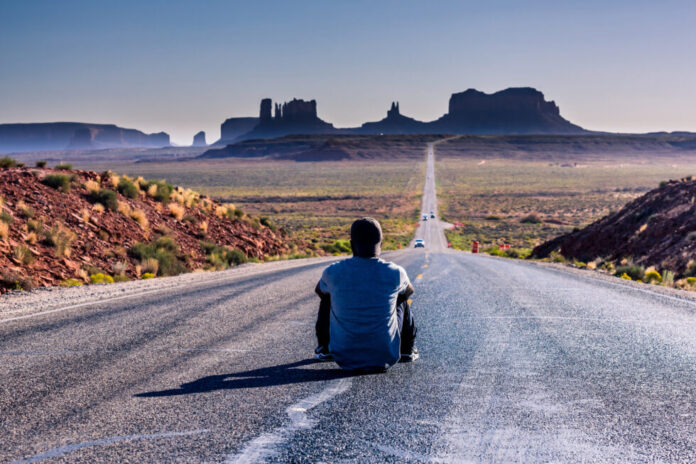 chłopak siedzi na środku drogi patrząc na monument valley w usa