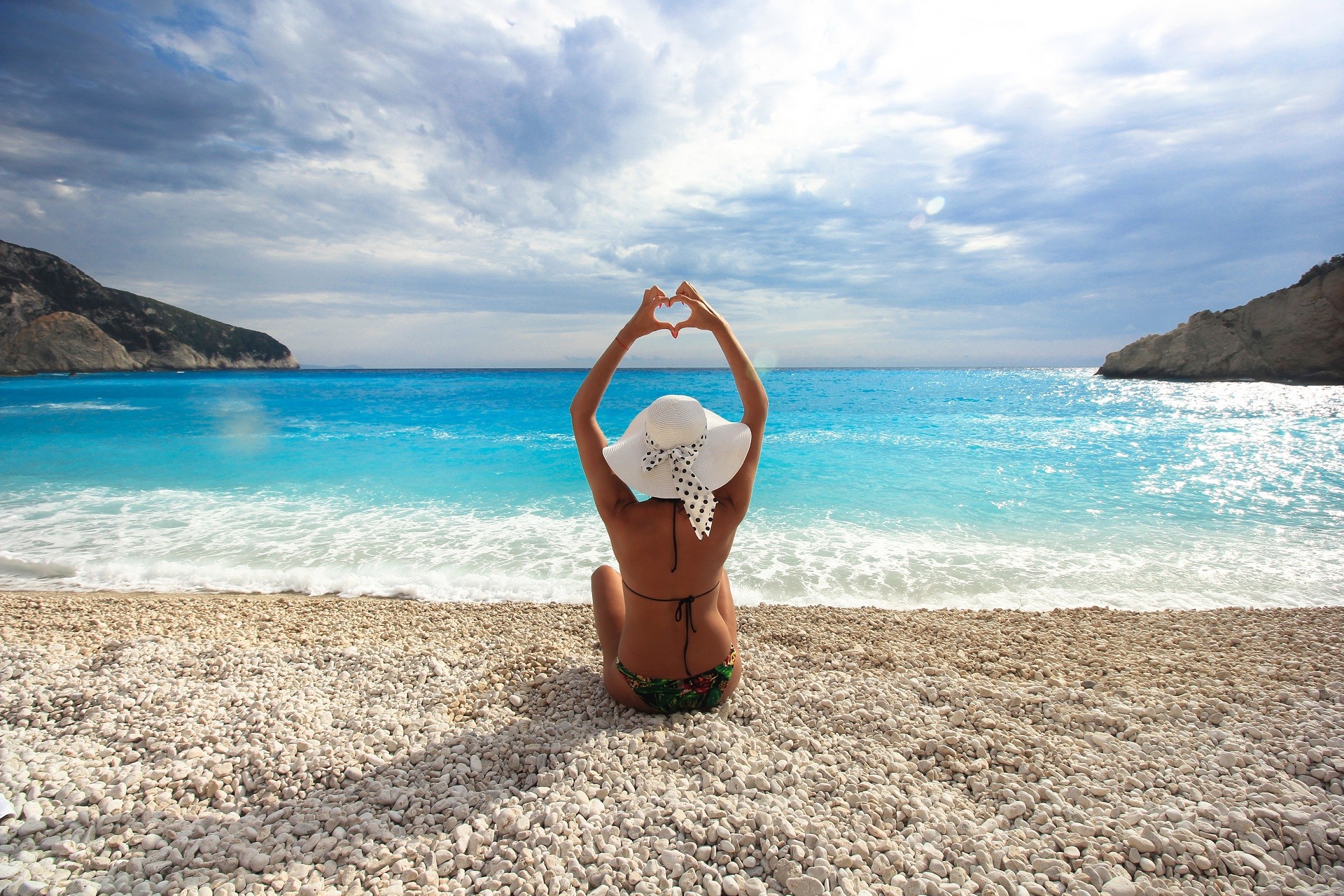 atrakcyjna kobieta siedząca na białej kamienistej plaży wykonująca gest dłońmi gest serca na tle turkusowego morza i błękitnego nieba