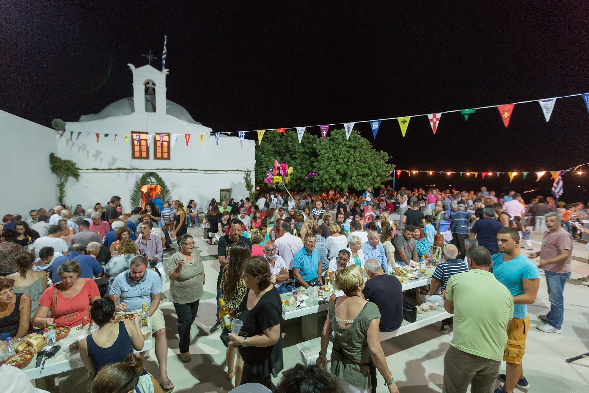 ludzie pijący i jedzący przy stołach przed białym kościołem udekorowanym kolorowymi proporcami