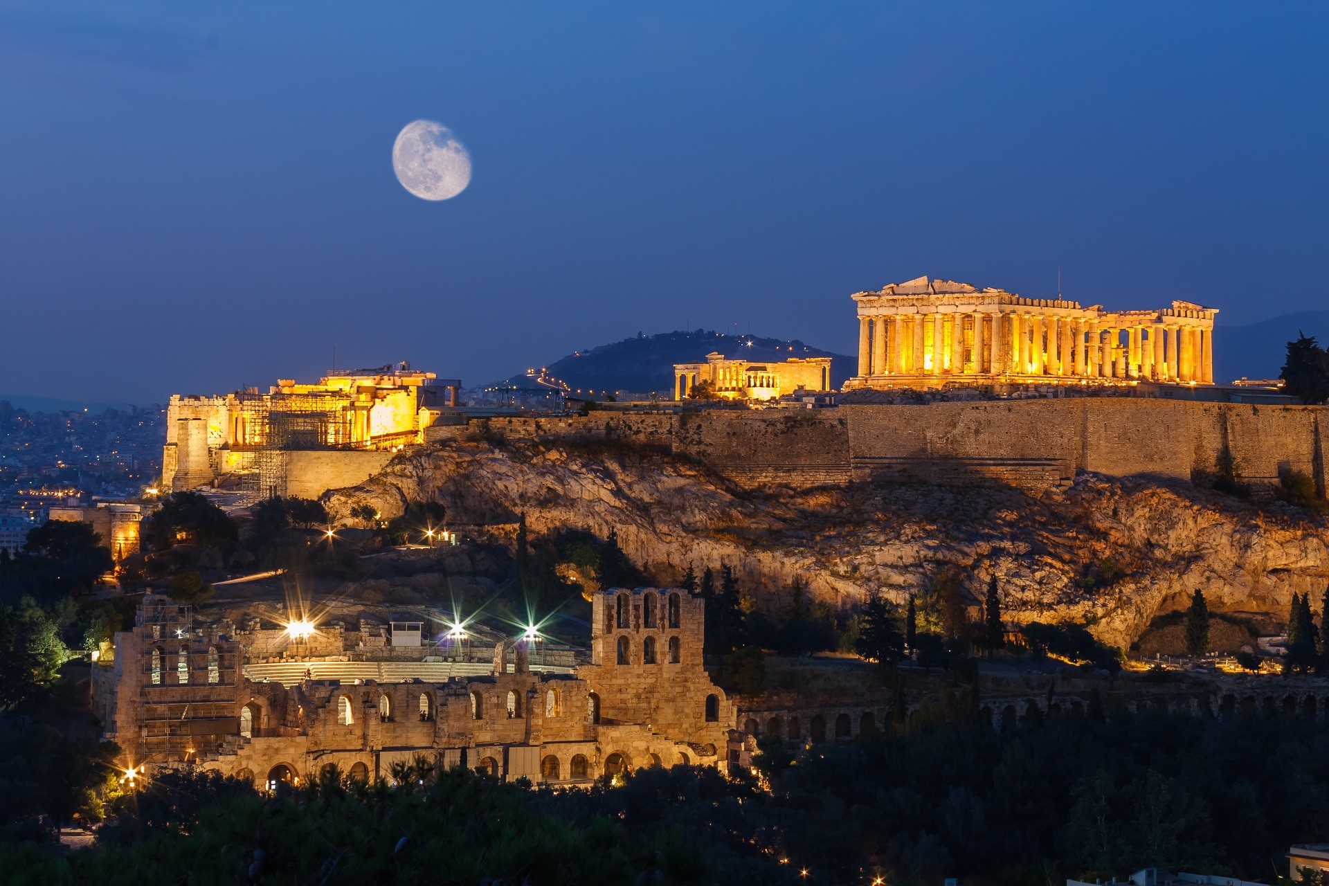 widok na podświetlone wzgórze Akropolu nocą na tle ciemnoniebieskiego nieba z księżycem