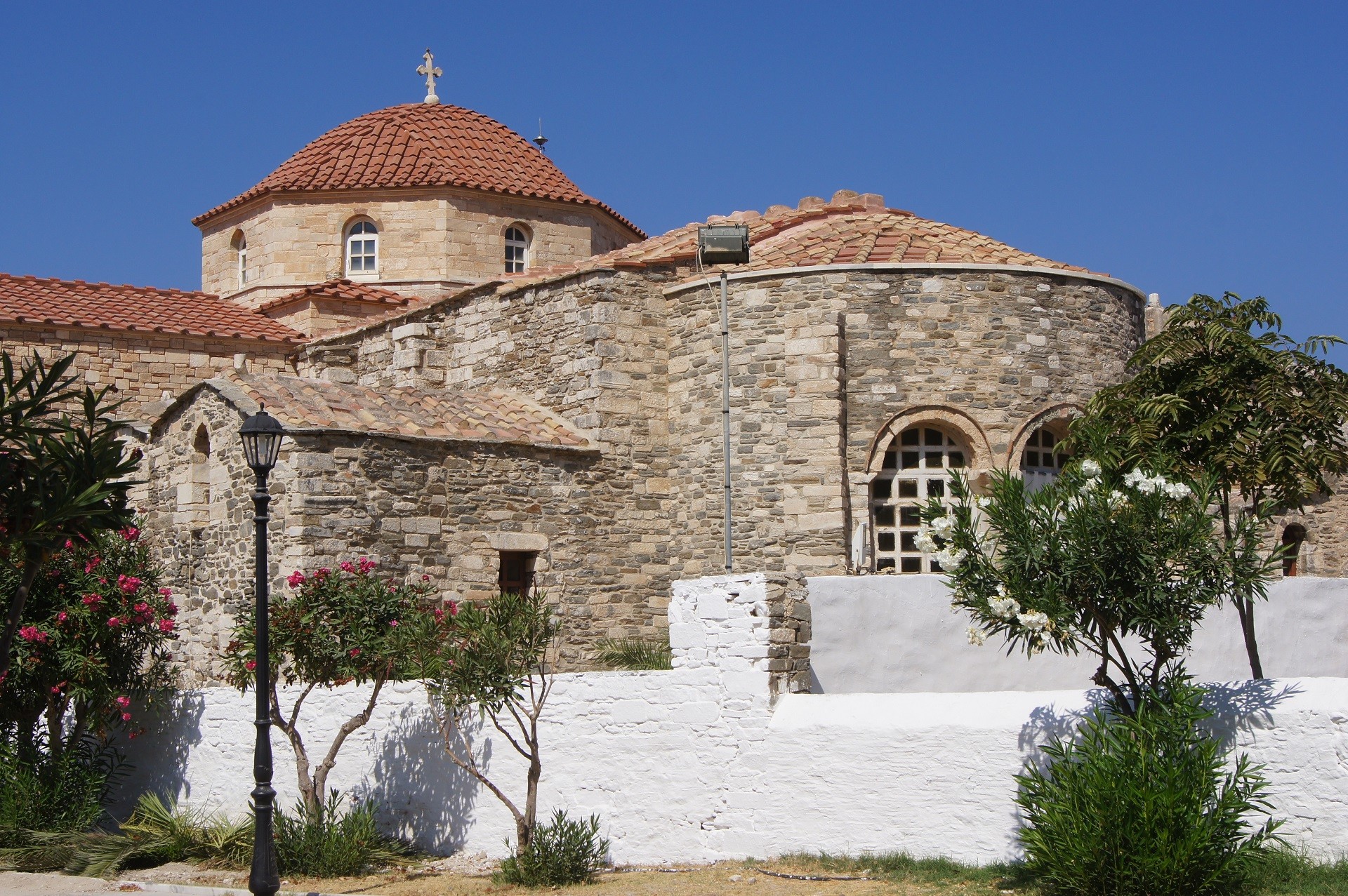 kościół wybudowany z kamienia w piaskowym kolorze z czerwonymi kopułami na tle niebieskiego nieba, przed kościołem biały niski murek