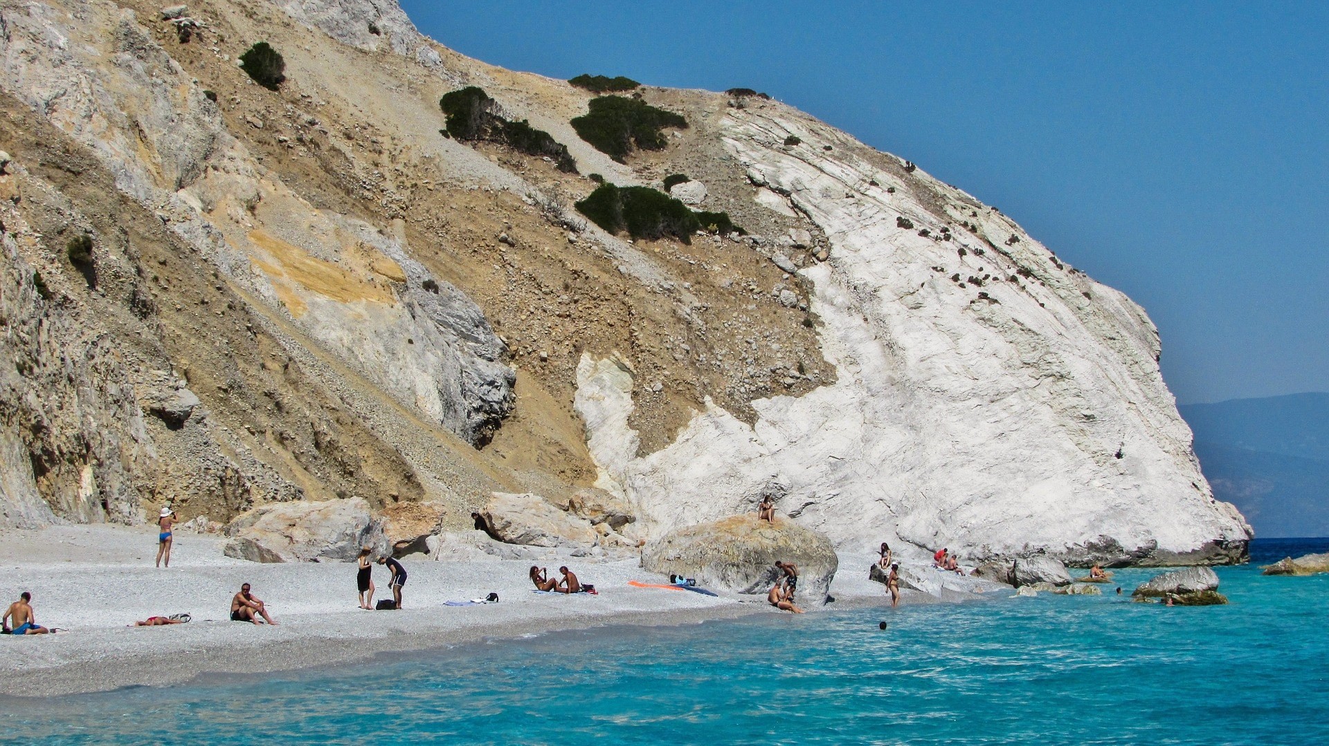 kamienista biała plaża oblana turkusowymi wodami, na której wypoczywają ludzie na tle biało-kawowego wzgórza